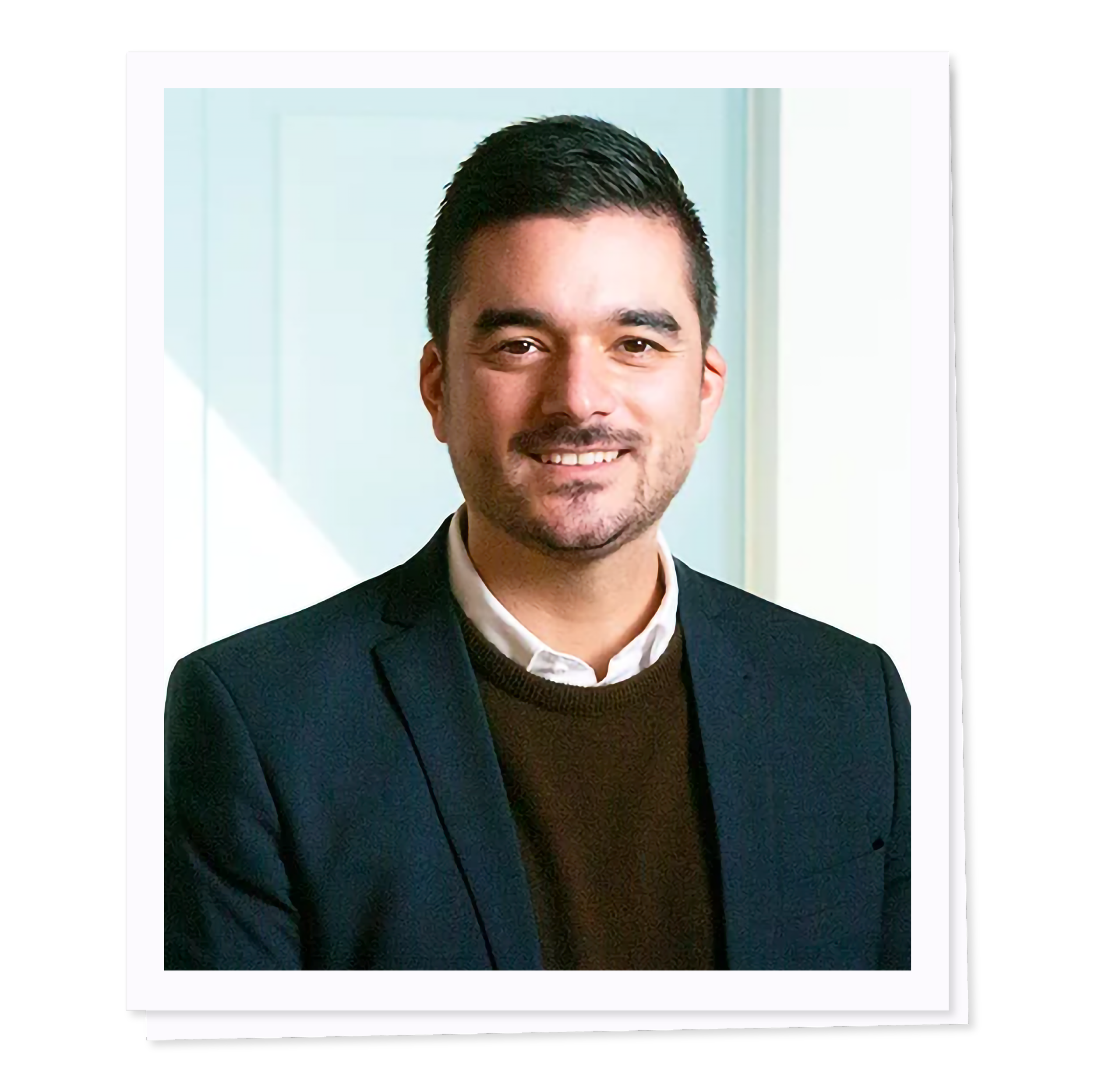 A man in a dark suit and brown sweater, smiling, with short hair, standing in front of a light-colored background.