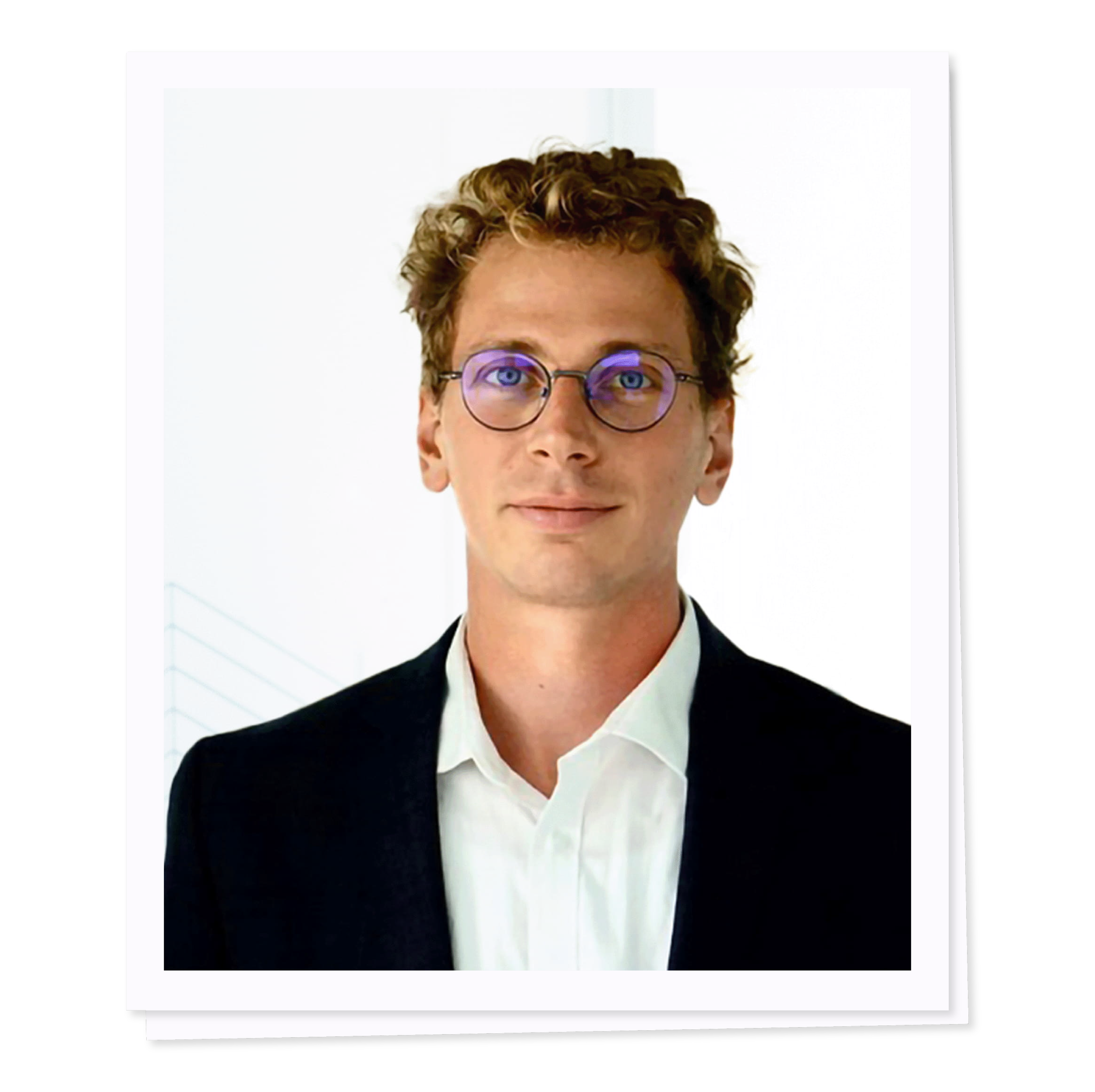 A man with wavy hair, wearing round glasses, a white shirt, and a dark blazer, against a plain background.