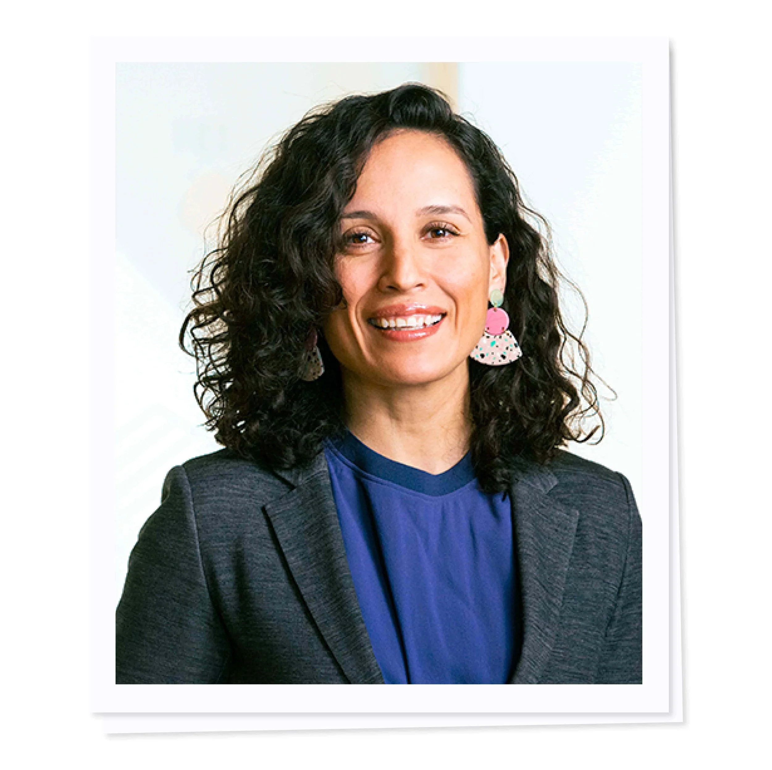 A woman with curly hair smiling, wearing colorful earrings, a blue blouse, and a gray blazer.