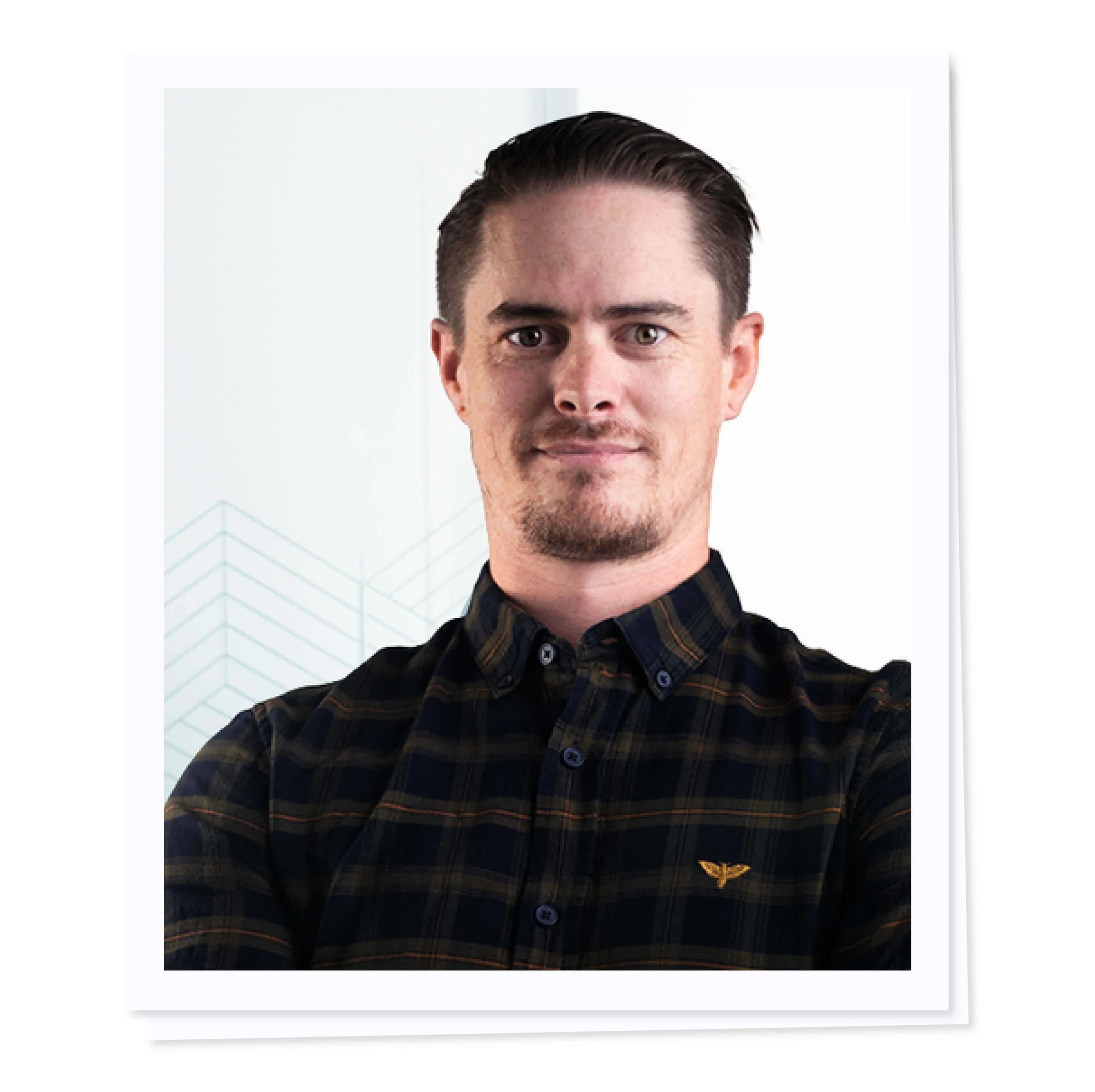 A man with short hair and a goatee wearing a plaid button-up shirt, standing against a light background.