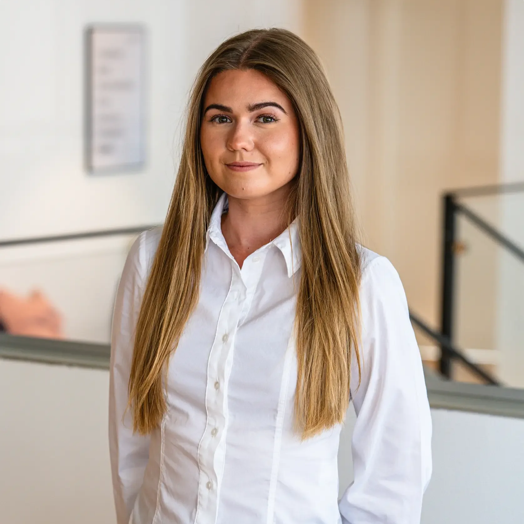 Smiling woman with long hair, wearing a black top and pearl necklace, standing against a light background.