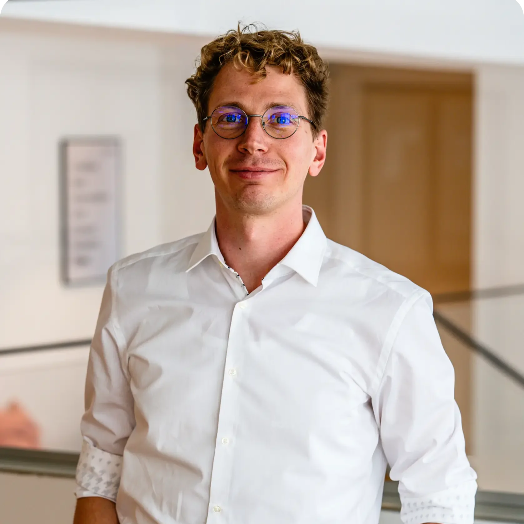 A man with curly hair, wearing glasses and a formal suit, is looking at the camera with a neutral expression. The background is plain white.
