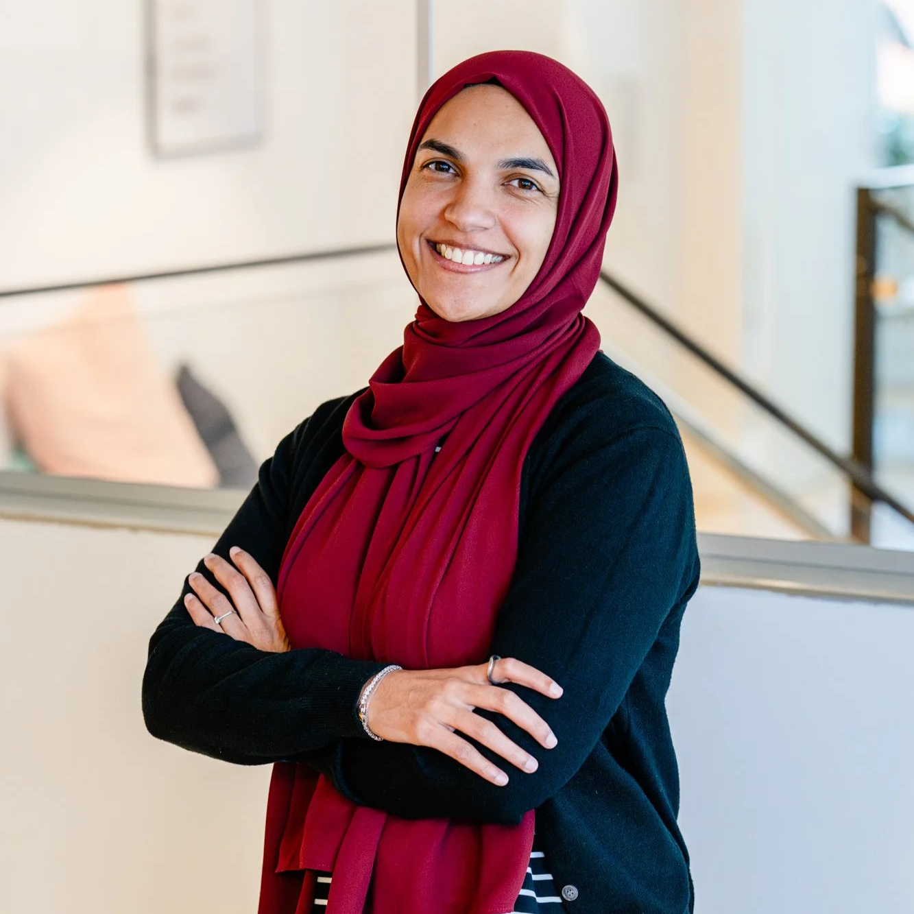 Smiling person wearing a light pink hijab and a maroon top, standing against a light background.