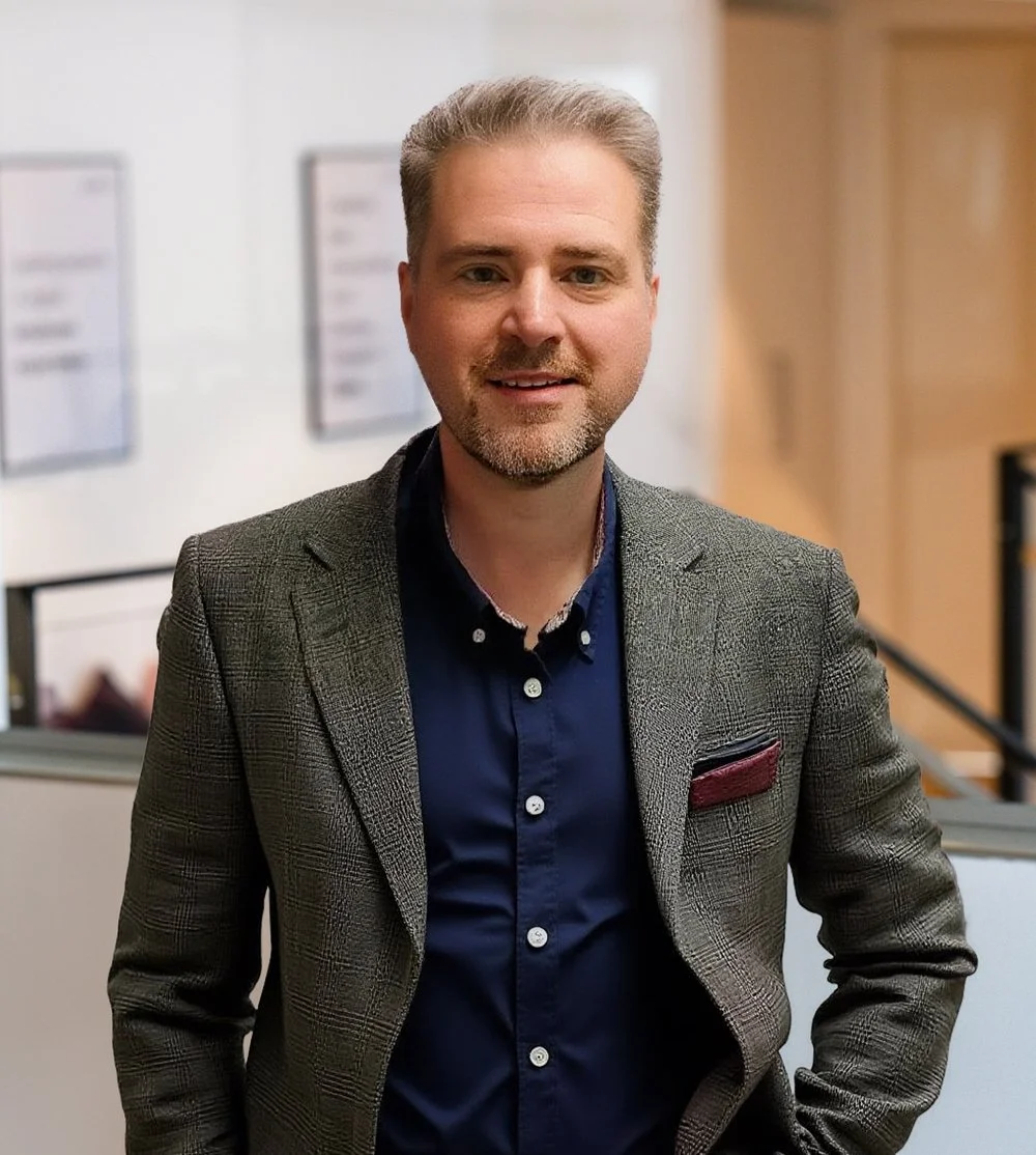 A smiling man in a gray blazer and navy shirt with a light beard.