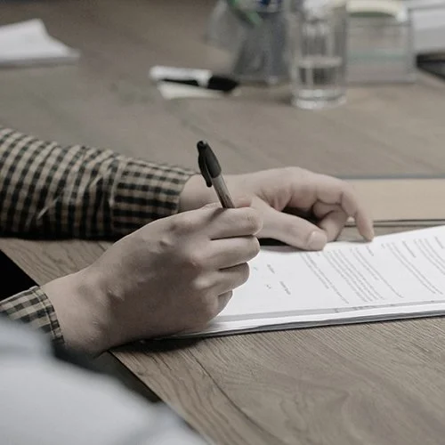 Person wearing a plaid shirt signing or writing on a document with a black pen on a wooden table.