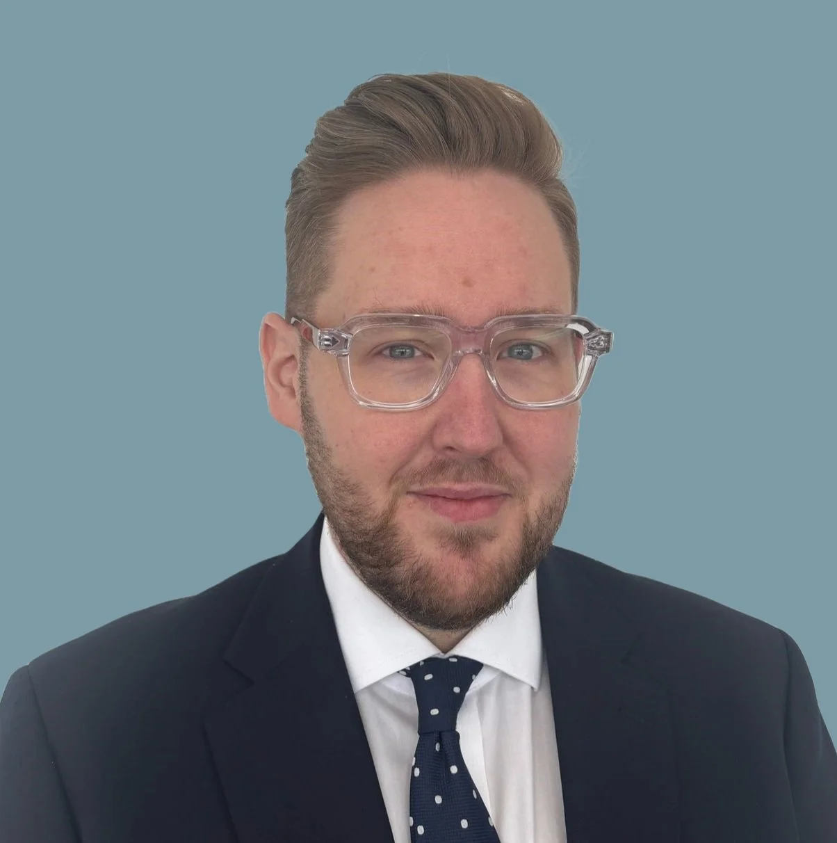 Portrait of a man with glasses, wearing a suit and polka dot tie, against a light blue background.