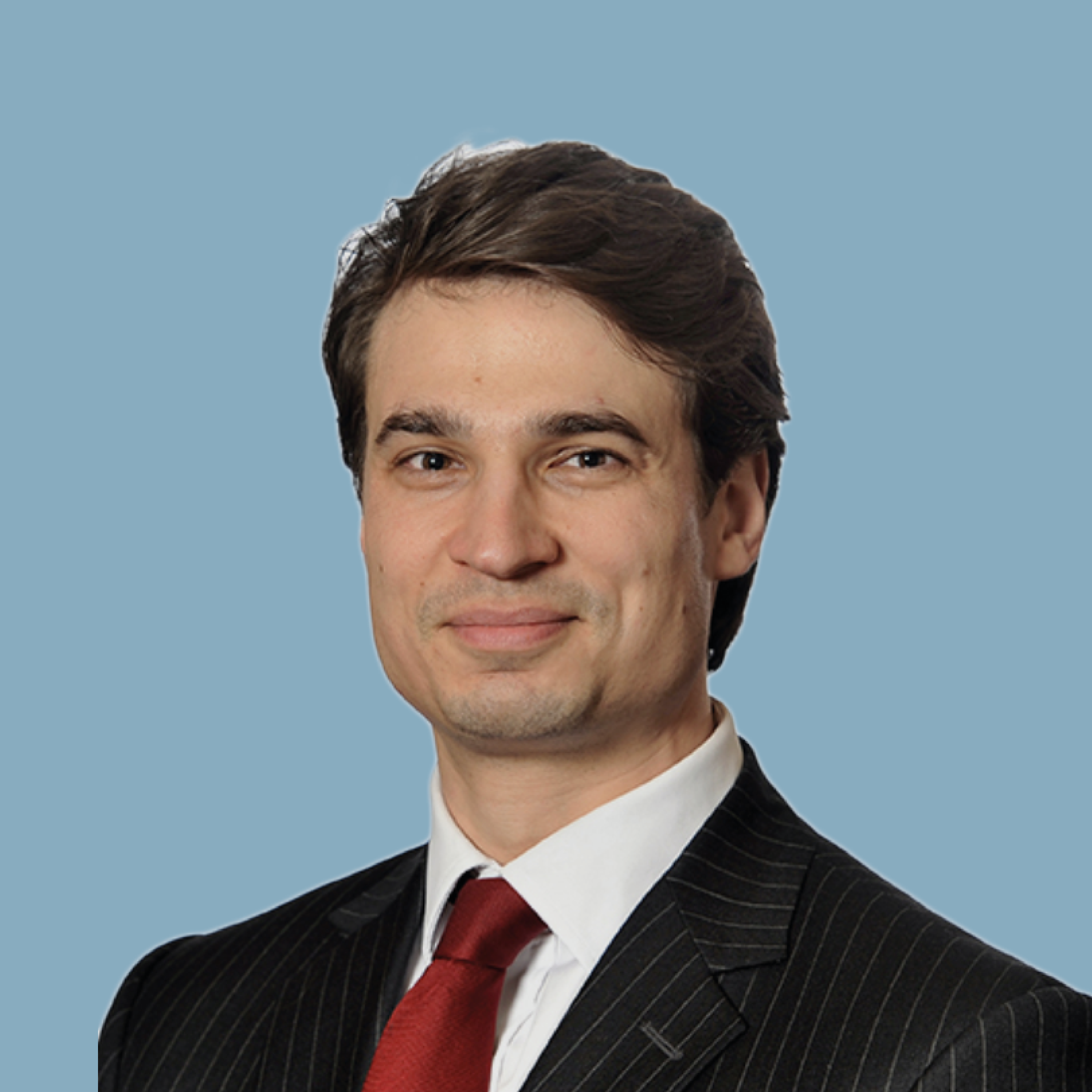 Close-up of a man with brown hair wearing a black pinstripe suit, white shirt, and red tie, against a blue background.