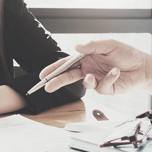 Close-up of a handshake between two people, one holding a pen, with documents on a wooden table.