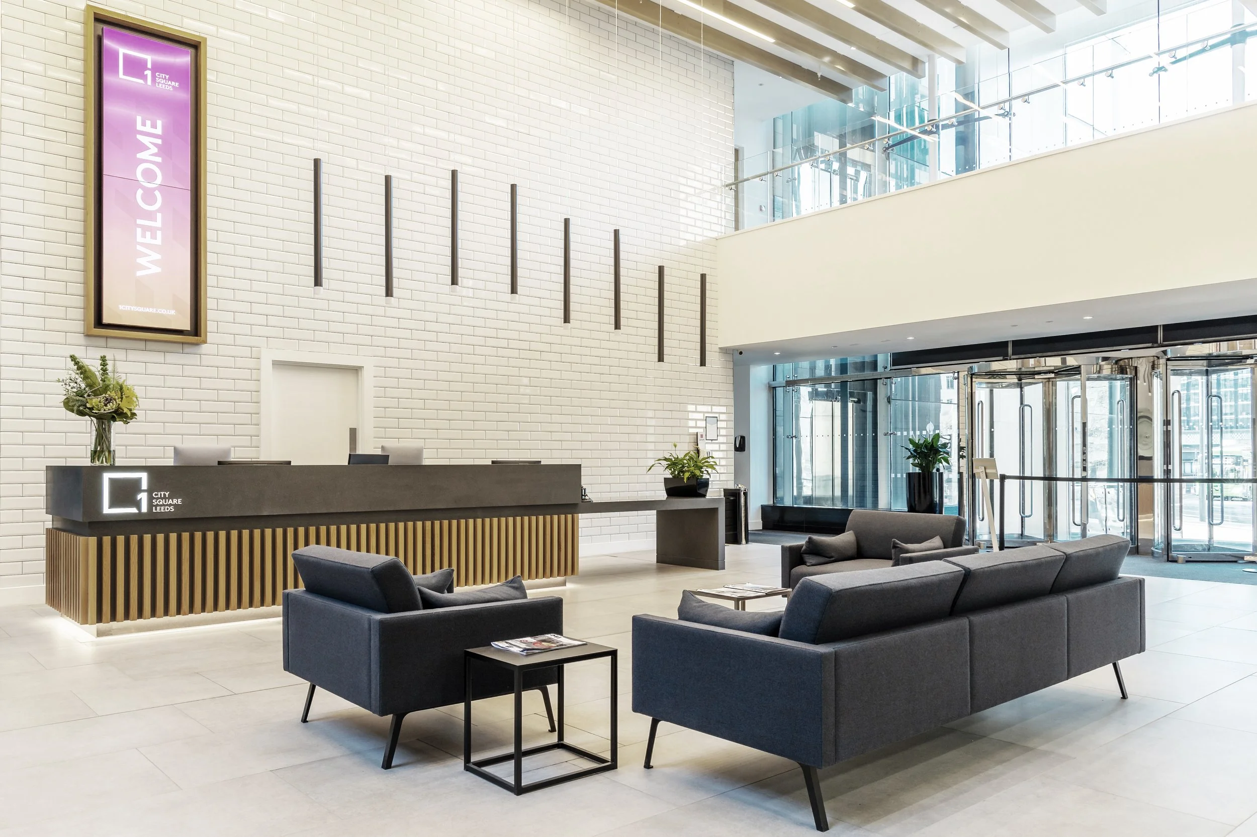 Modern hotel lobby with a front desk, gray sofas, black chairs, large windows, plants, and a welcome sign for City Square Leeds.
