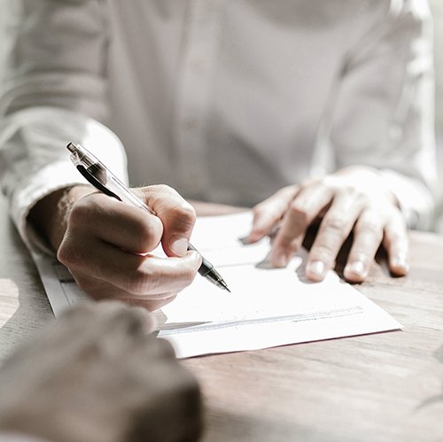 Person writing with a pen on a piece of paper on a wooden table.