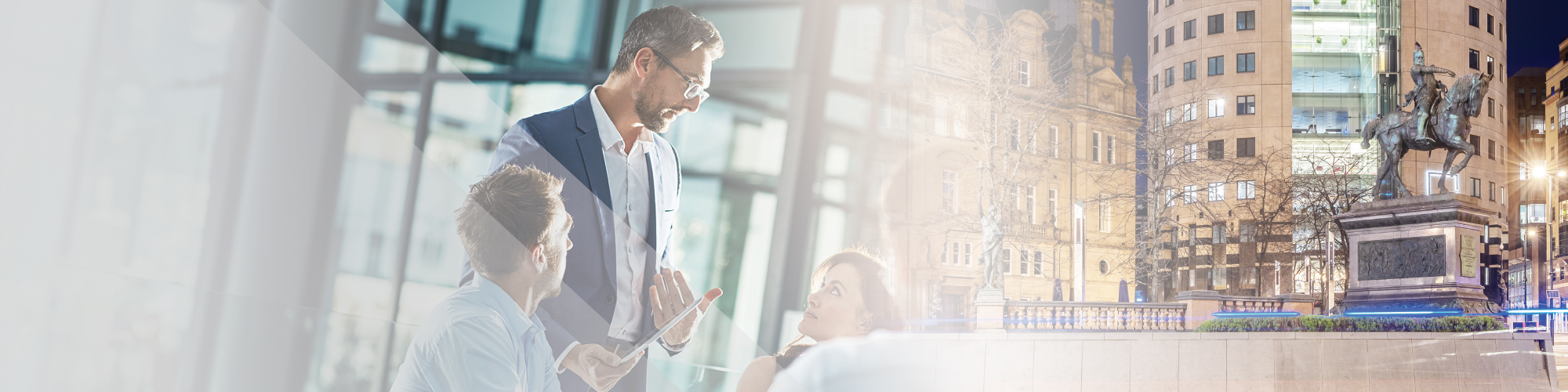 Two men and one woman in a modern office with large glass windows, one man standing and the other seated talking to a woman, city buildings and monuments visible through the windows at night.