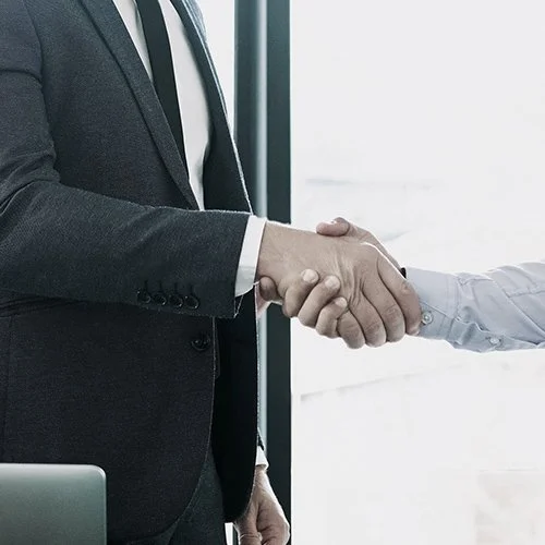 Close-up of two people shaking hands, one in a dark suit and the other in a light-colored shirt, near a window in a professional setting.