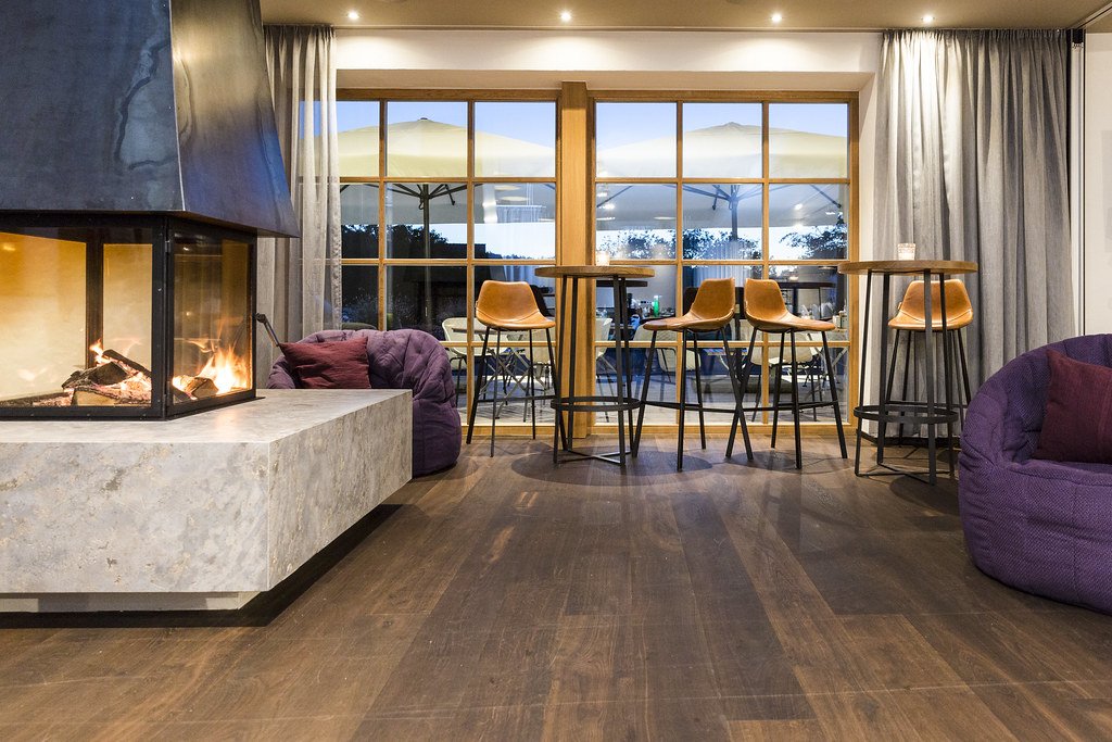 Gemütliches Wohnzimmer mit Kamin, bequemen Sitzen, hohem Tisch, Barhockern, großen Fenstern und Blick nach draußen auf eine Terrasse mit Sonnenschirmen