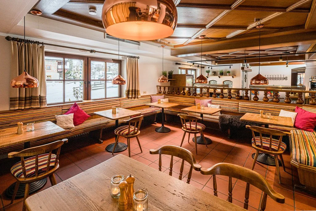 Ein gemütliches Restaurant mit Holztischen, Stühlen und bunten Sitzbänken. Es gibt Wand- und Deckenleuchten im Copper-Look, große Fenster mit Vorhängen und Dekor auf der Fensterbank. Das Interieur hat eine warme, einladende Atmosphäre.