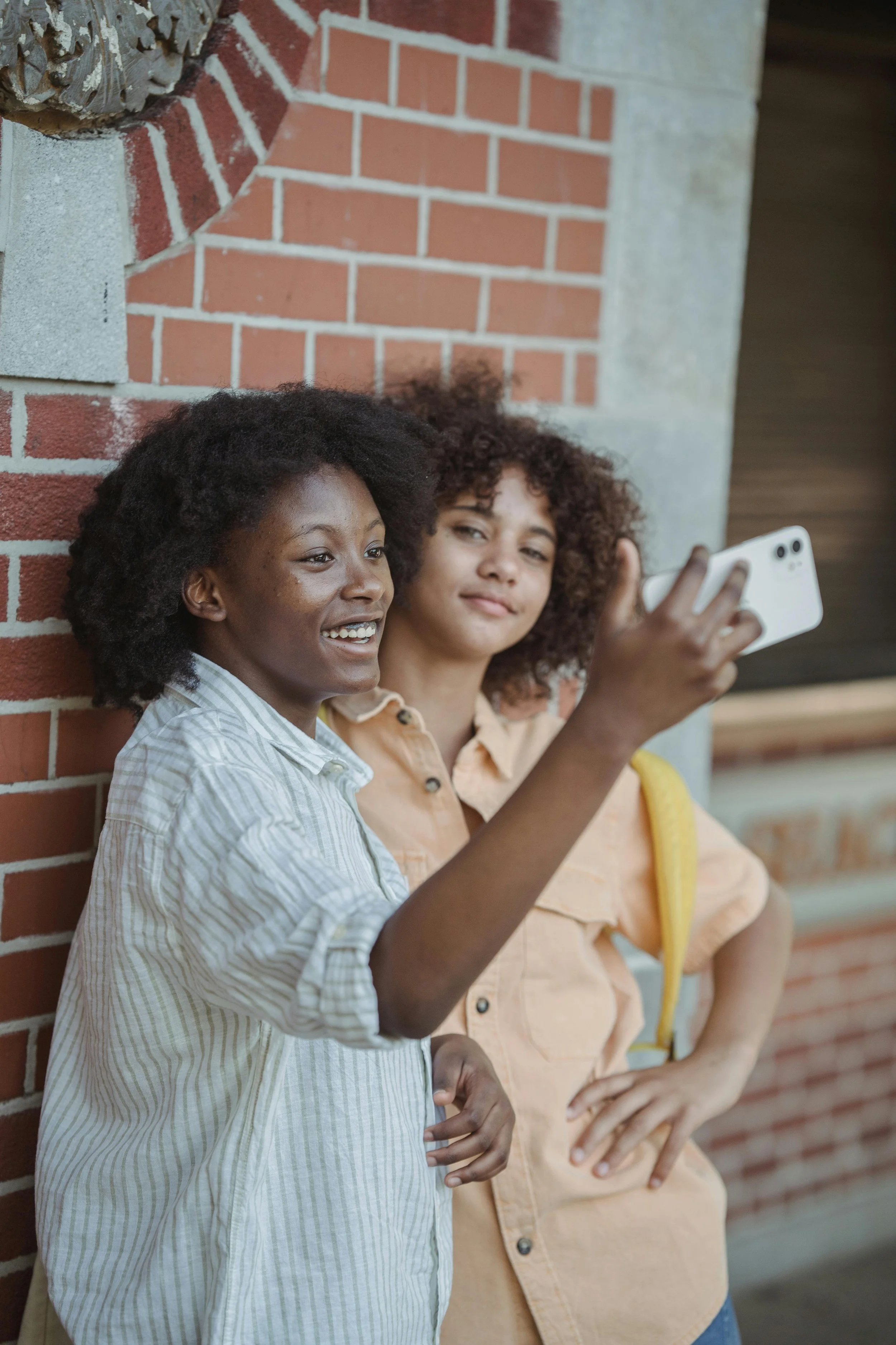 Zwei Frauen sitzen an einer roten Backsteinwand, eine nimmt ein Selfie auf.