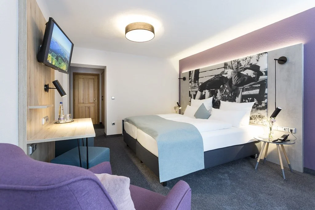 Modernes Hotelzimmer mit Doppelbett, Wandkunst, Fernseher, Schreibtisch mit Wasserflasche und Gläser, Leseleuchten, gemütlicher Sessel, Weiche Farbgebung.