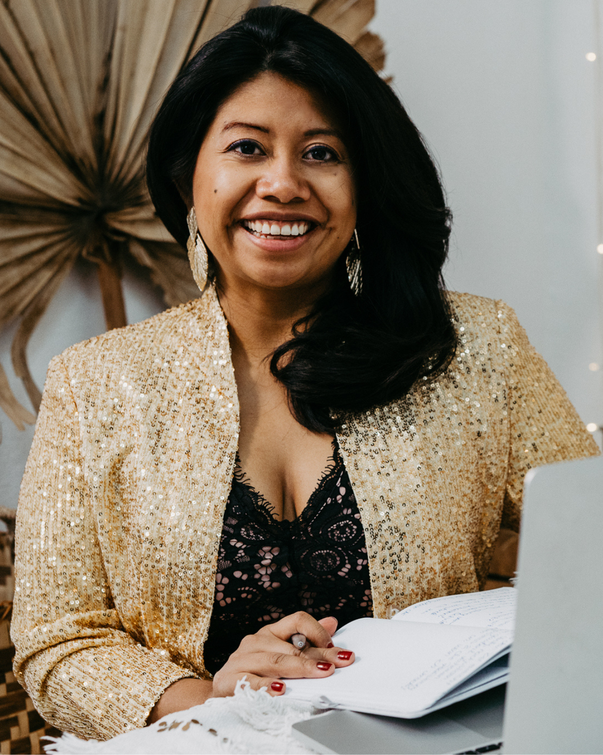 Frau mit schwarzen Haaren, goldener Paillettenkleid und schwarzer Spitzenbluse sitzt vor einem Laptop, lächelt und schaut in die Kamera.