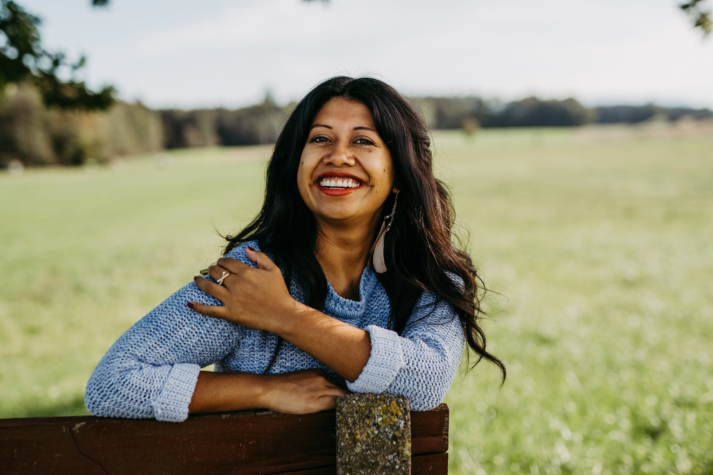 Frau sitzt lachend auf einer Parkbank unter einem Baum in einem offenen Feld.