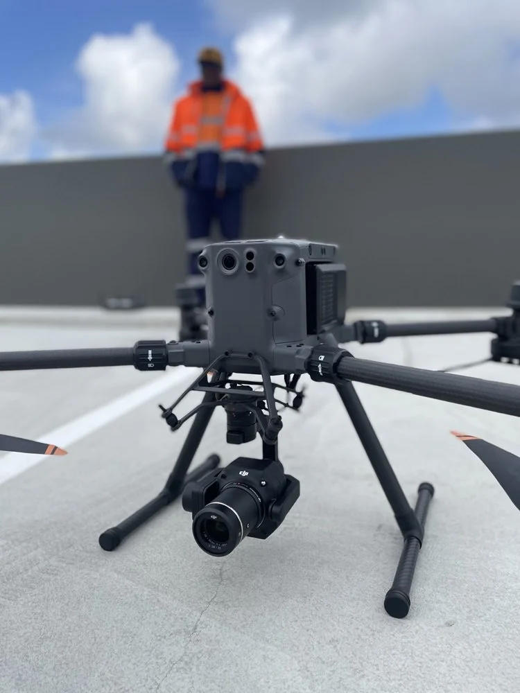 A drone with a mounted camera on a rooftop, with a person in safety gear standing in the background against a cloudy sky.
