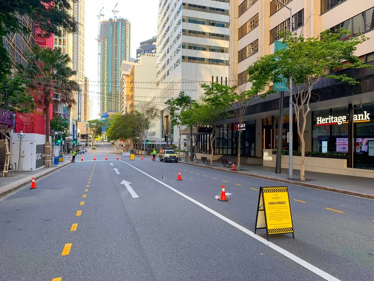 City street with road closure signs and cones, workers in safety vests, tall buildings, and trees along the sidewalk.