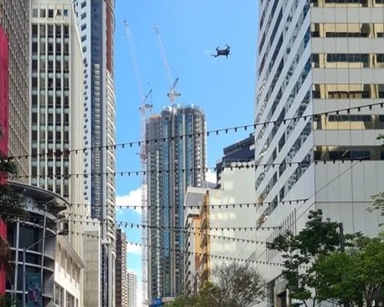 A helicopter flying over city skyscrapers with construction cranes on tall buildings and string lights hanging across the street.