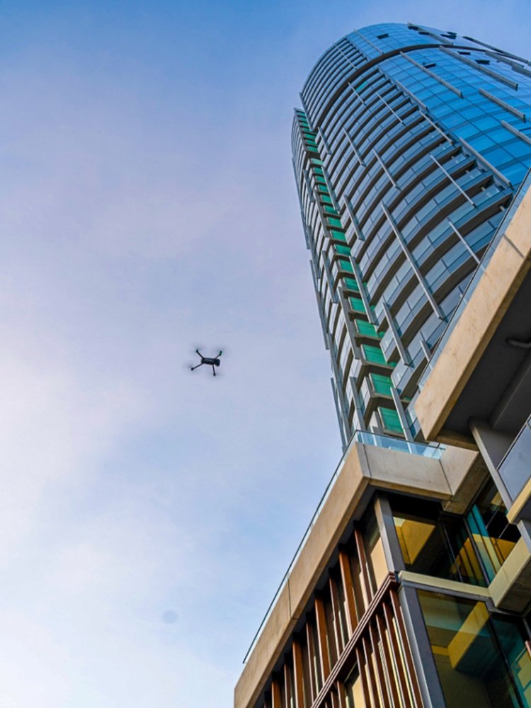 A tall modern skyscraper with glass windows and balconies, viewed from below with a drone flying in the sky nearby.