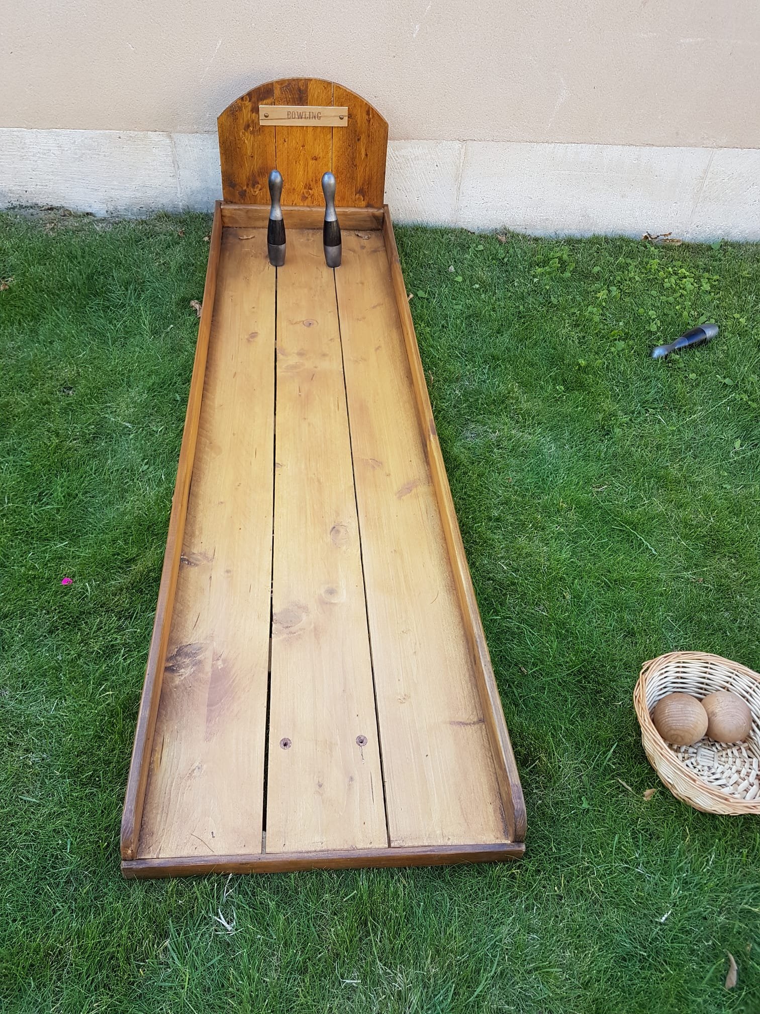 Jeu de bowling en bois avec deux quilles, une balle et un panier avec des balles sur l'herbe près d'un mur.