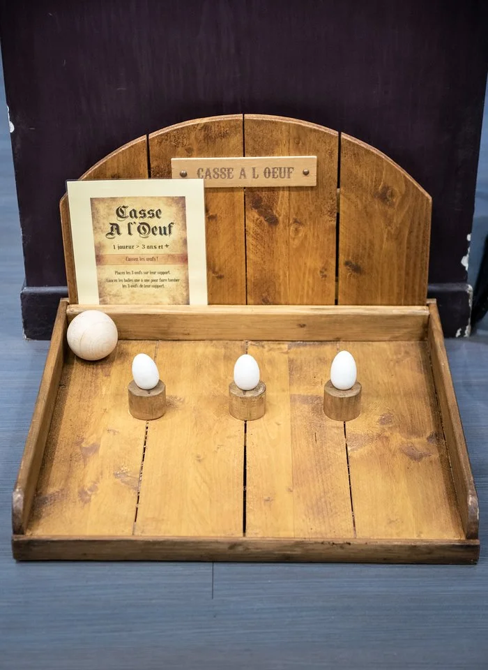 Jeu en bois pour casser des œufs, avec trois œufs blancs placés sur des supports en bois, et un panneau indiquant 'Casse à l'œuf', dans un lieu intérieur.