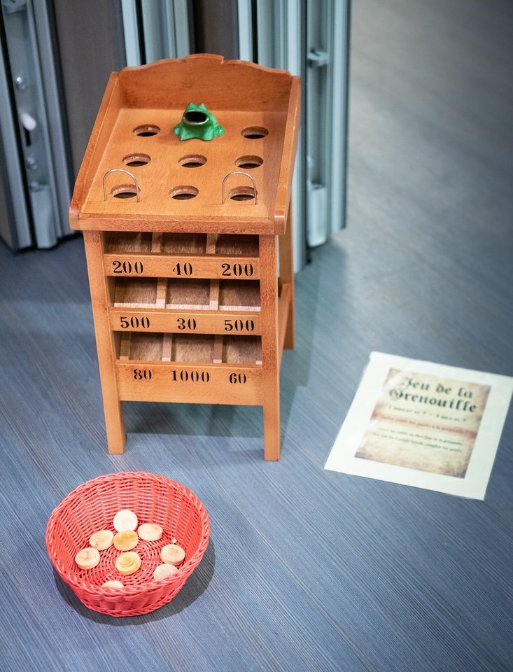 Une table en bois pour le jeu de la grenouille, avec des trous sur le dessus et un petit animal en plastique vert. Au sol, un panier en rotin rouge contenant des jetons en bois. Une affiche dans le fond indique 'Jeu de la Grenouille'.