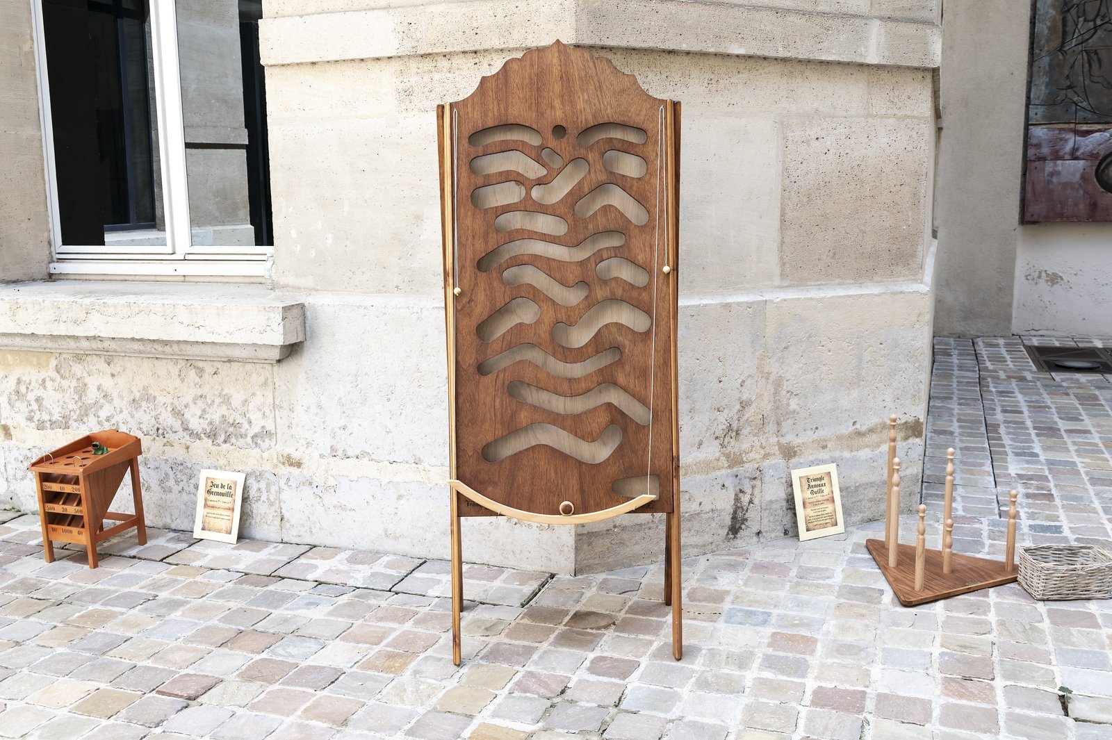 Installation artistique en bois posée sur une allée pavée, comprenant une pièce centrale avec des formes ondulées découpées, un petit meuble avec des tiroirs, une affiche et un support à broches en bois.