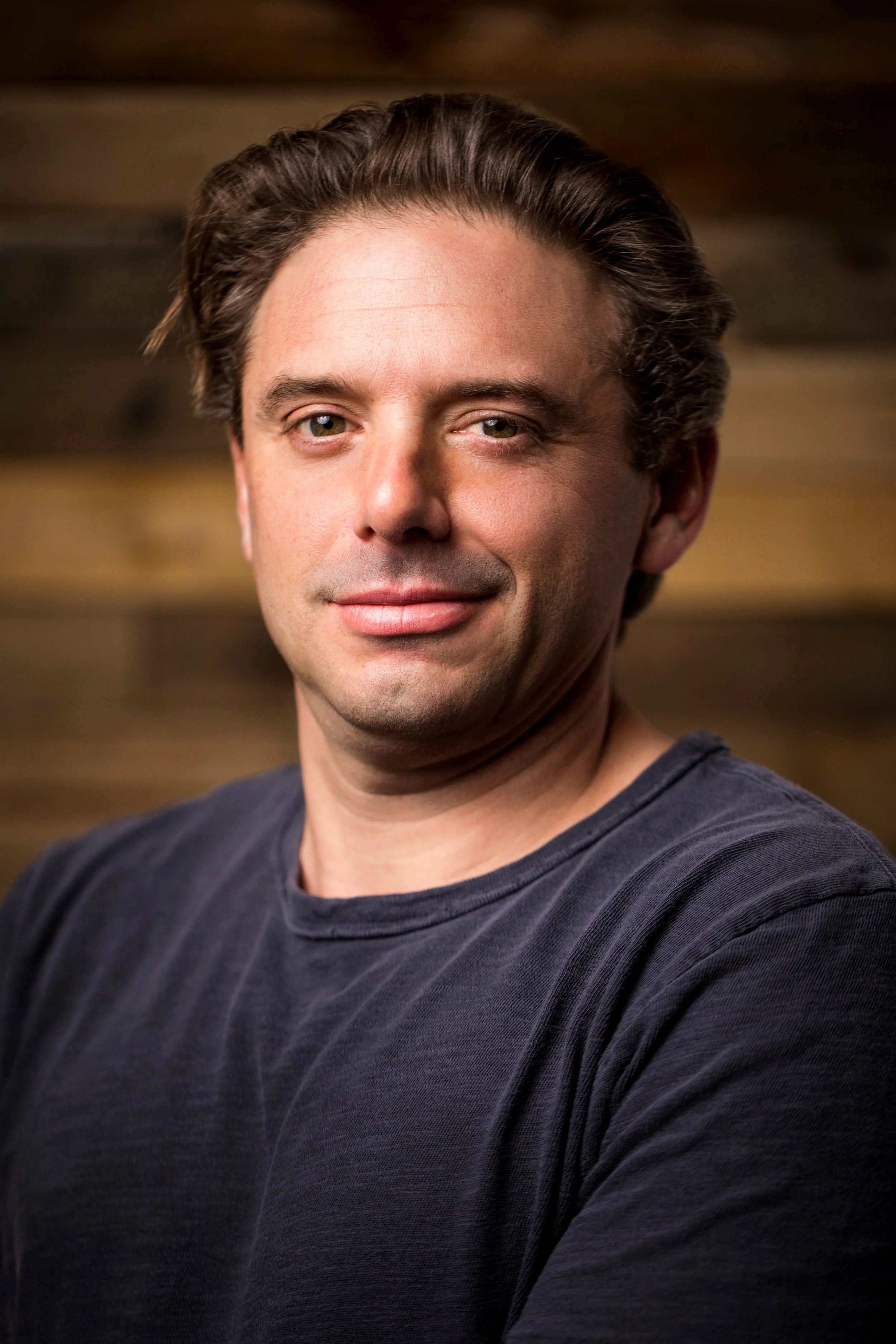 Close-up portrait of a man with dark hair smiling, wearing a dark shirt, against a blurred wooden background.