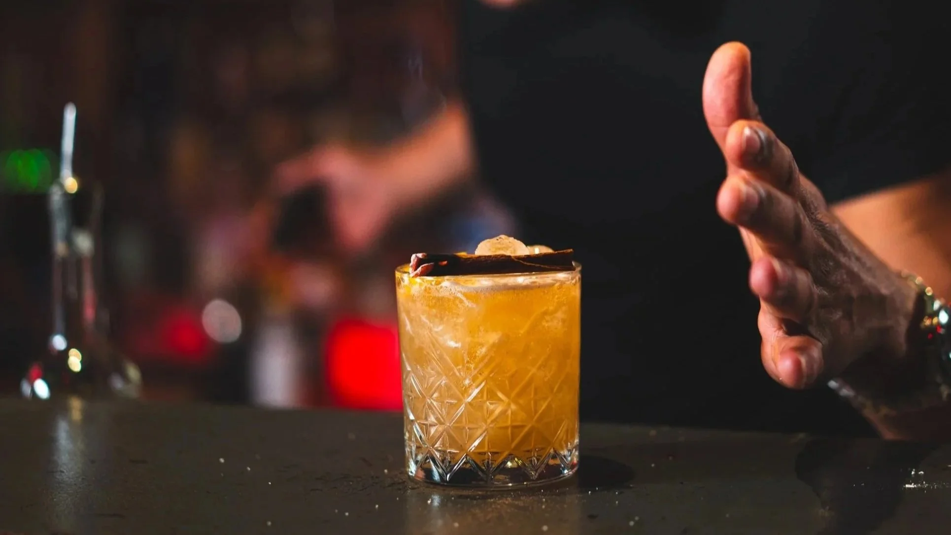 A cocktail with ice in a textured glass, garnished with a piece of chocolate and a small round fruit, on a dark bar counter. A bartender's hand is nearby, preparing to serve.