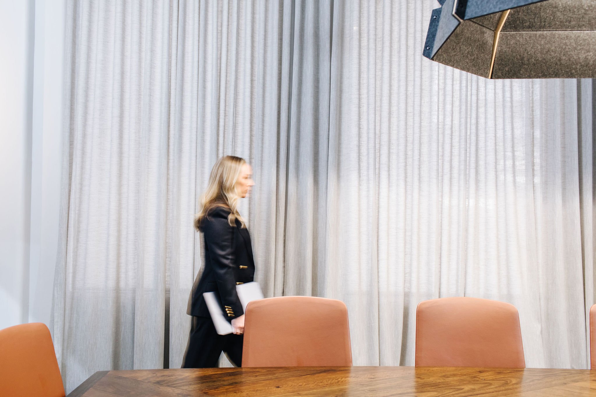 A woman in a black blazer and pants walking through a conference room with a wooden table and orange chairs, holding a laptop. Light-colored curtains are in the background.
