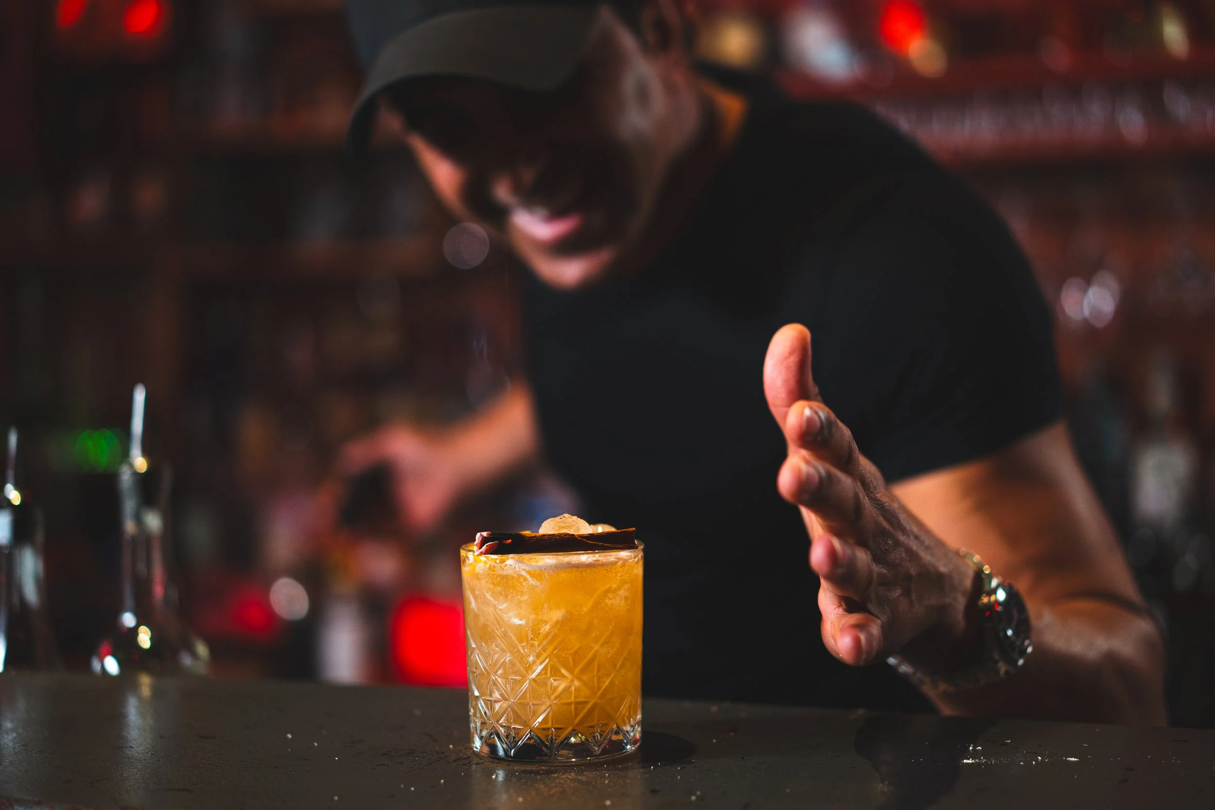 A bartender behind a bar, smiling and gesturing towards a yellow cocktail with a garnish on top, served in a patterned glass.