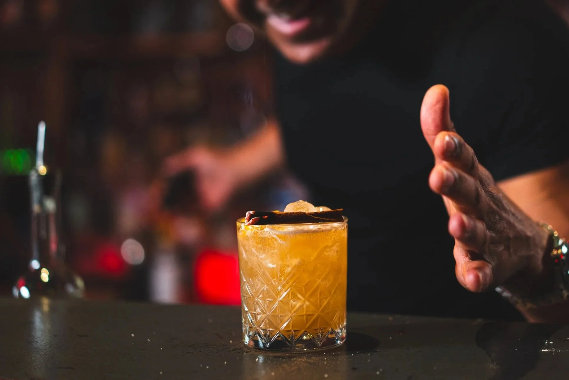 Close-up of a bartender preparing a cocktail in a short glass with ice, garnished with a piece of dark chocolate and a wafer cookie, on a bar counter.