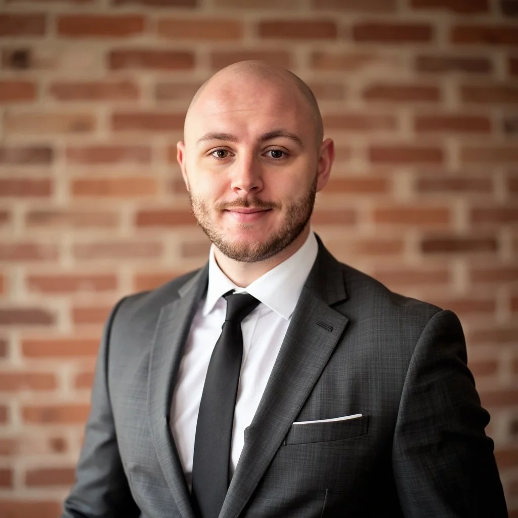 A man dressed in a dark gray suit, white shirt, and black tie standing in front of a brick wall.