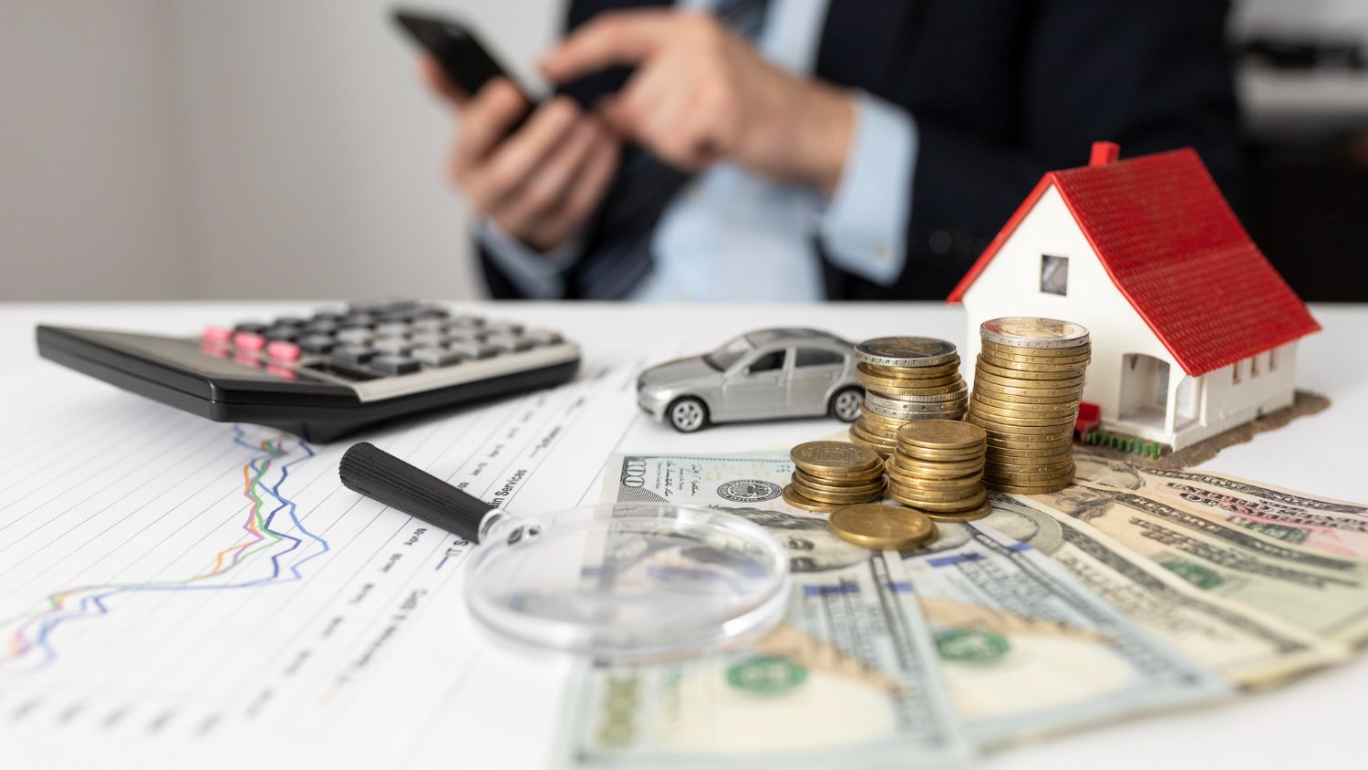 Financial planning scene with coins, dollar bills, a small model house, a toy car, a calculator, a magnifying glass, and a person using a smartphone in the background.