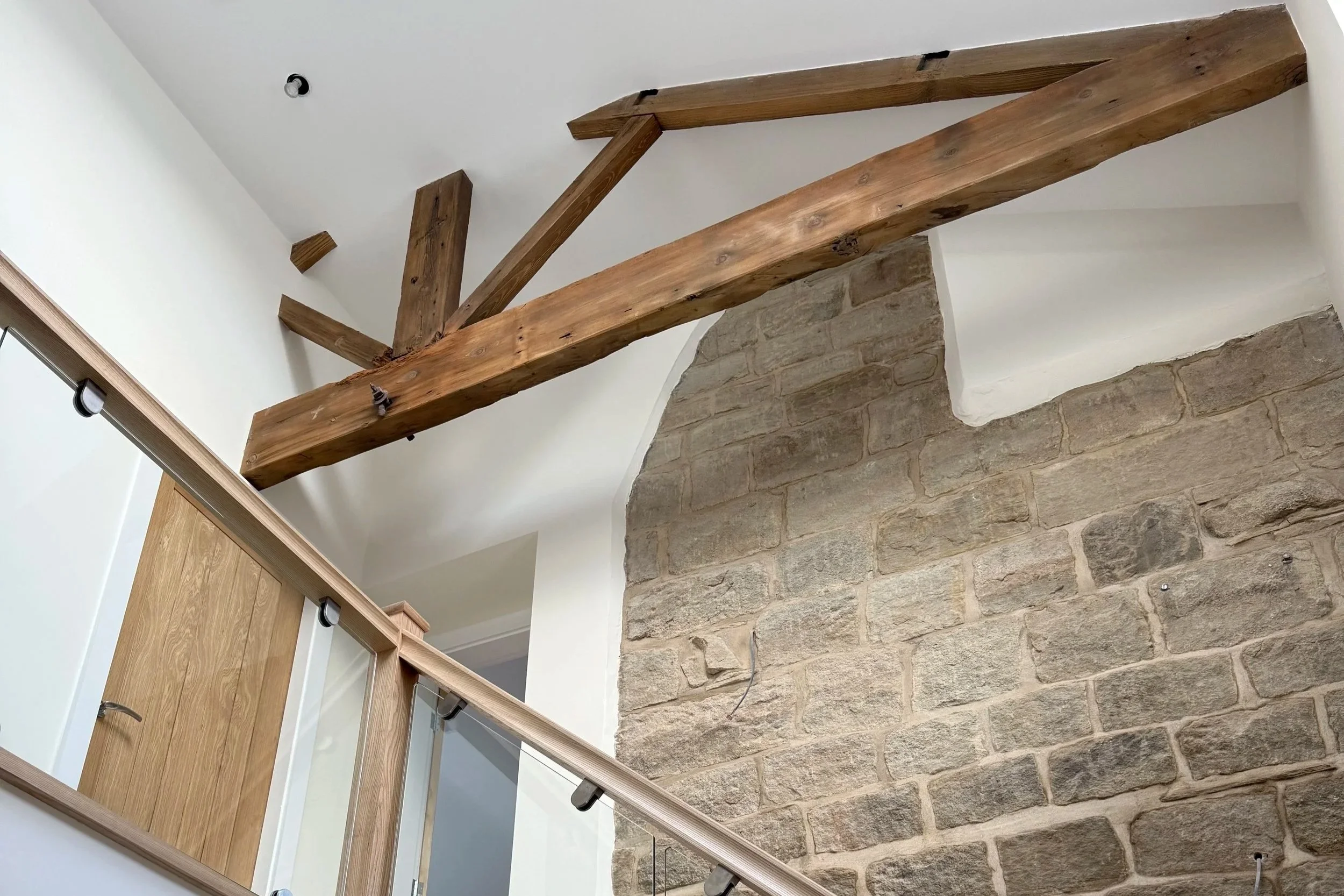 Interior view of a house with exposed wooden beams, white walls, a stone wall, and a wooden railing.