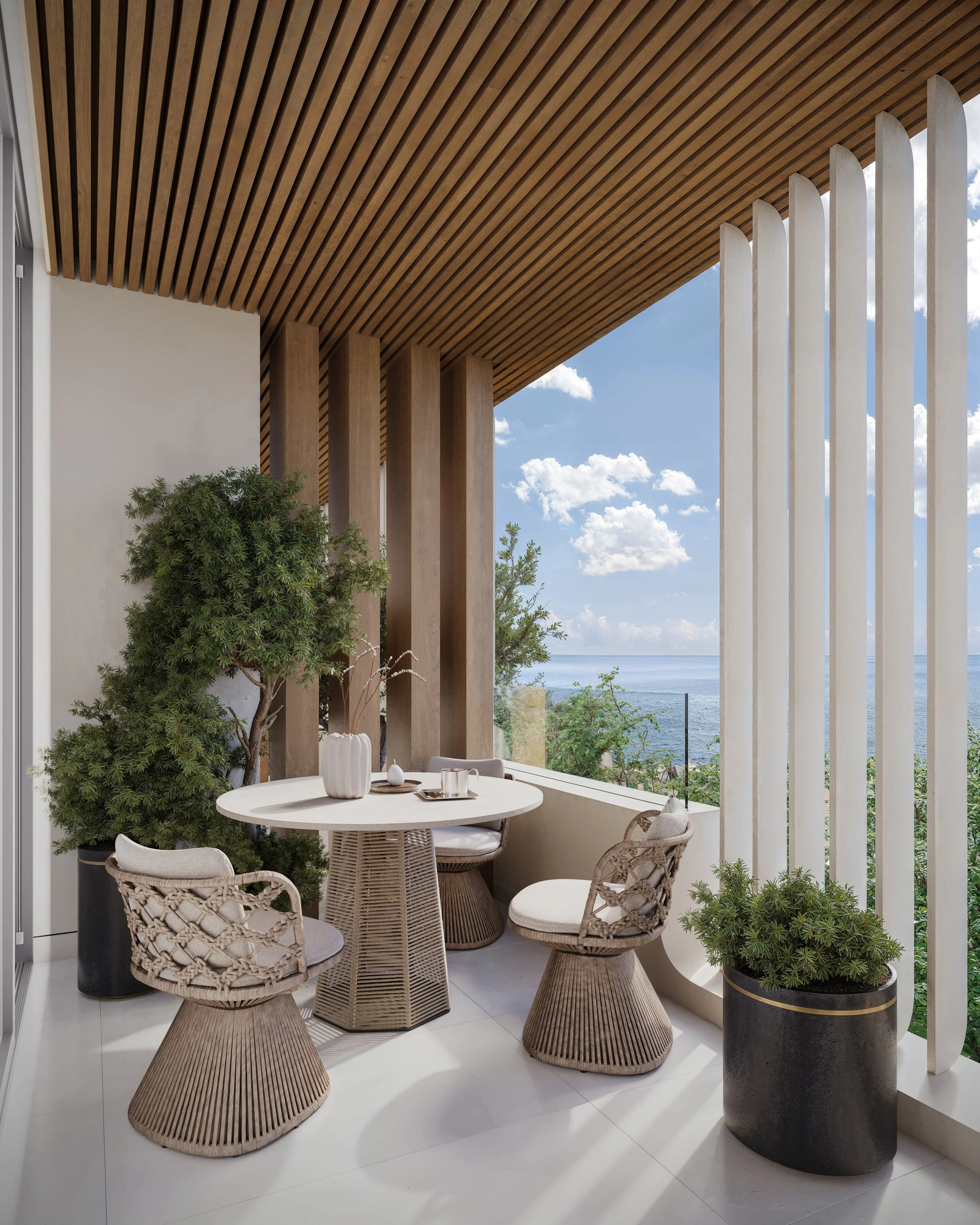 Balcon avec une table ronde, quatre chaises en osier, plantes vertes en pot et vue sur la mer en arrière-plan.