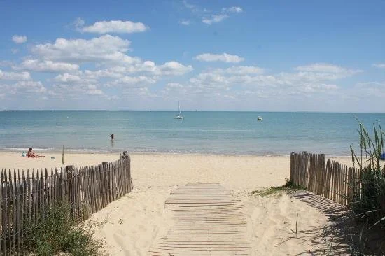 Wooden pathway leading to a sandy beach with calm ocean water and boats, bordered by wooden fences and grassy dunes under a partly cloudy sky.
