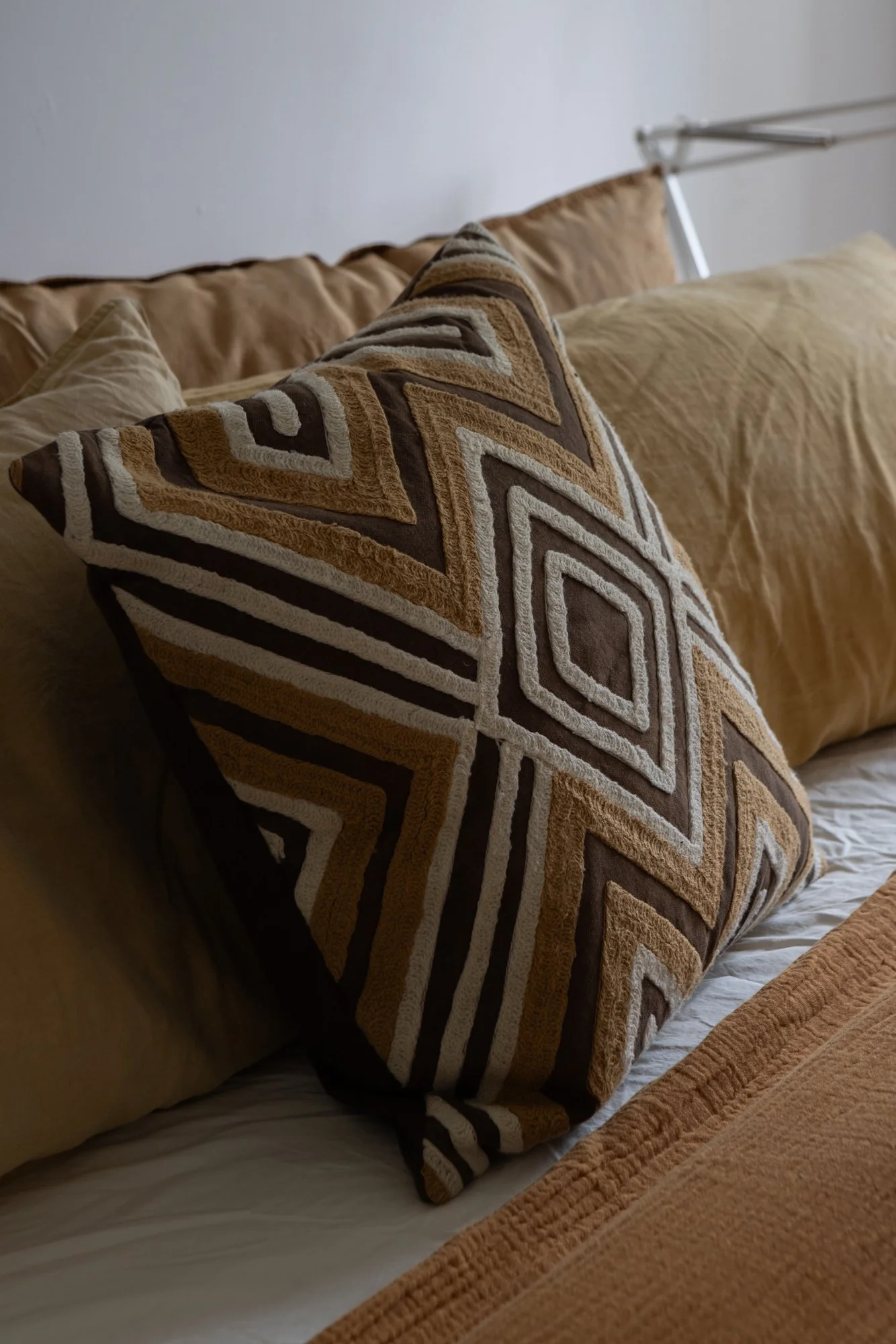 Close-up of decorative pillows on a bed, including a patterned pillow with geometric designs in shades of brown, beige, and white, and plain beige pillows behind it.