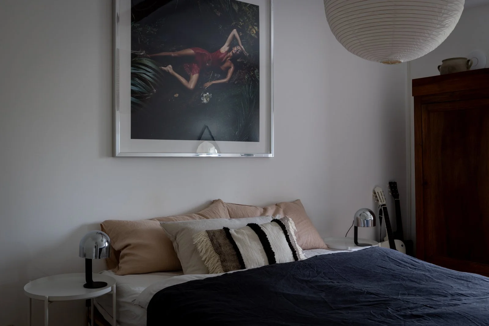 A bedroom with a bed that has beige and white pillows, a black duvet, and a white bed frame. On each side of the bed are round white nightstands with modern silver lamps. There is a large framed photograph above the bed of a woman lying on the ground