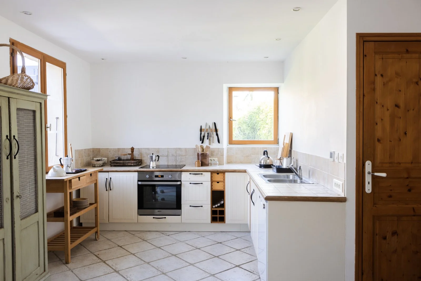 Kitchen with white cabinets, beige tiled countertops, stainless steel oven, wooden shelves, and a window showing greenery outside.