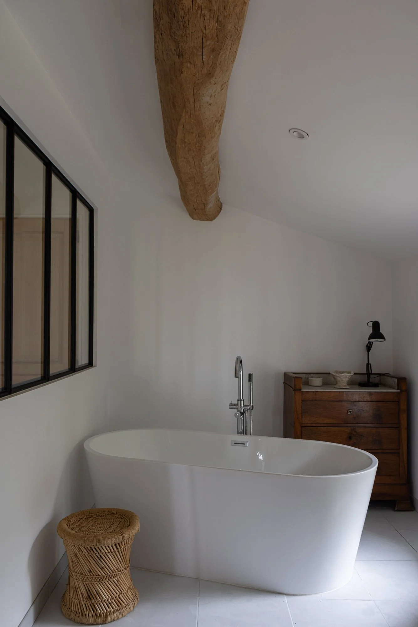 Modern bathroom with a free-standing white bathtub, wicker stool, wooden cabinet with accessories, black desk lamp, and a large wooden beam ceiling.