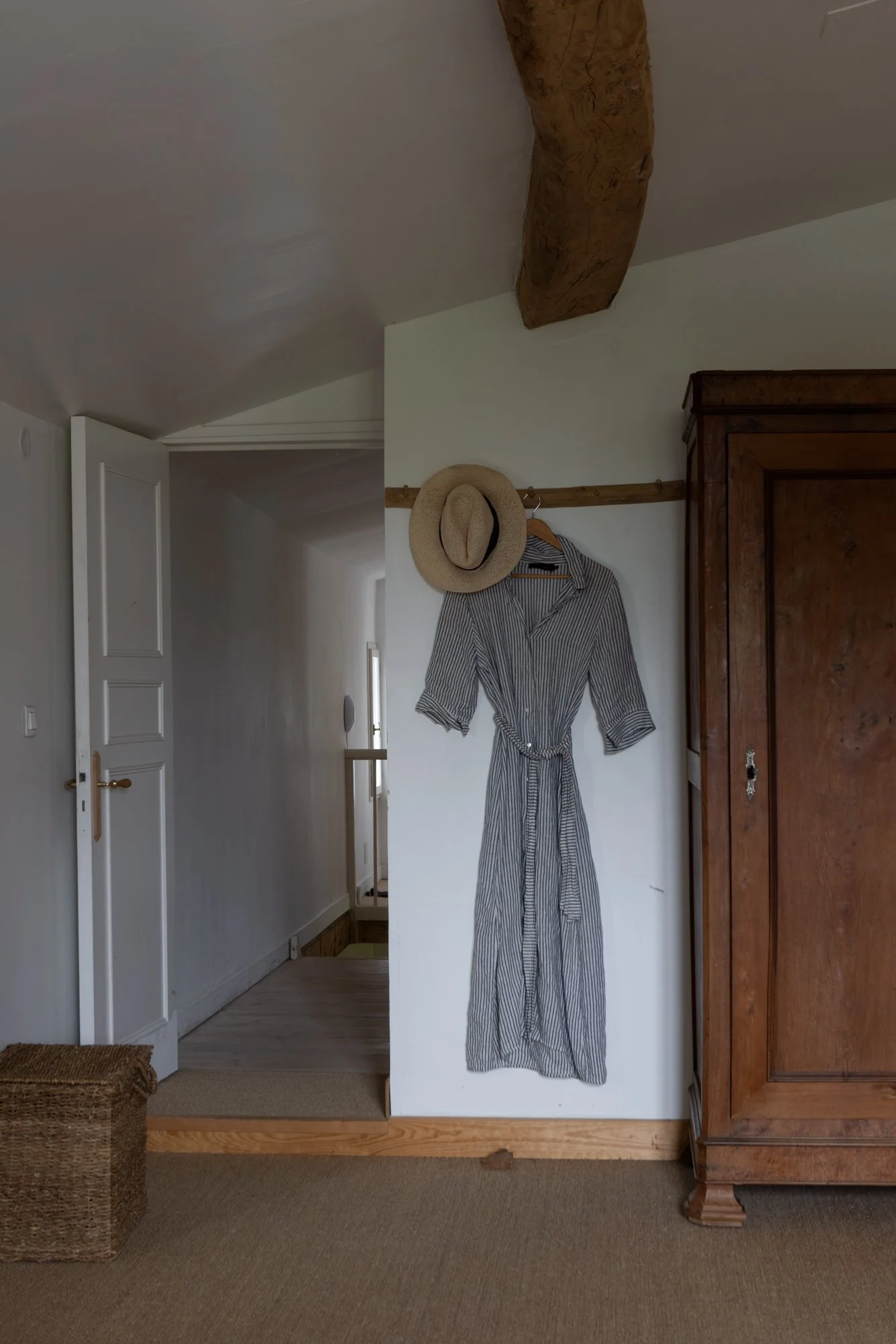 A striped dress hanging on a wooden hook on a white wall, with a straw hat hanging above it, next to a large wooden wardrobe.