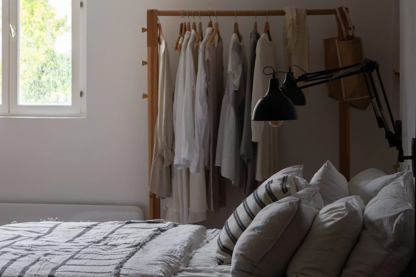 A cozy bedroom with a bed, pillows, and bed linens. There is a clothing rack with white and beige garments and a window with natural light coming in.