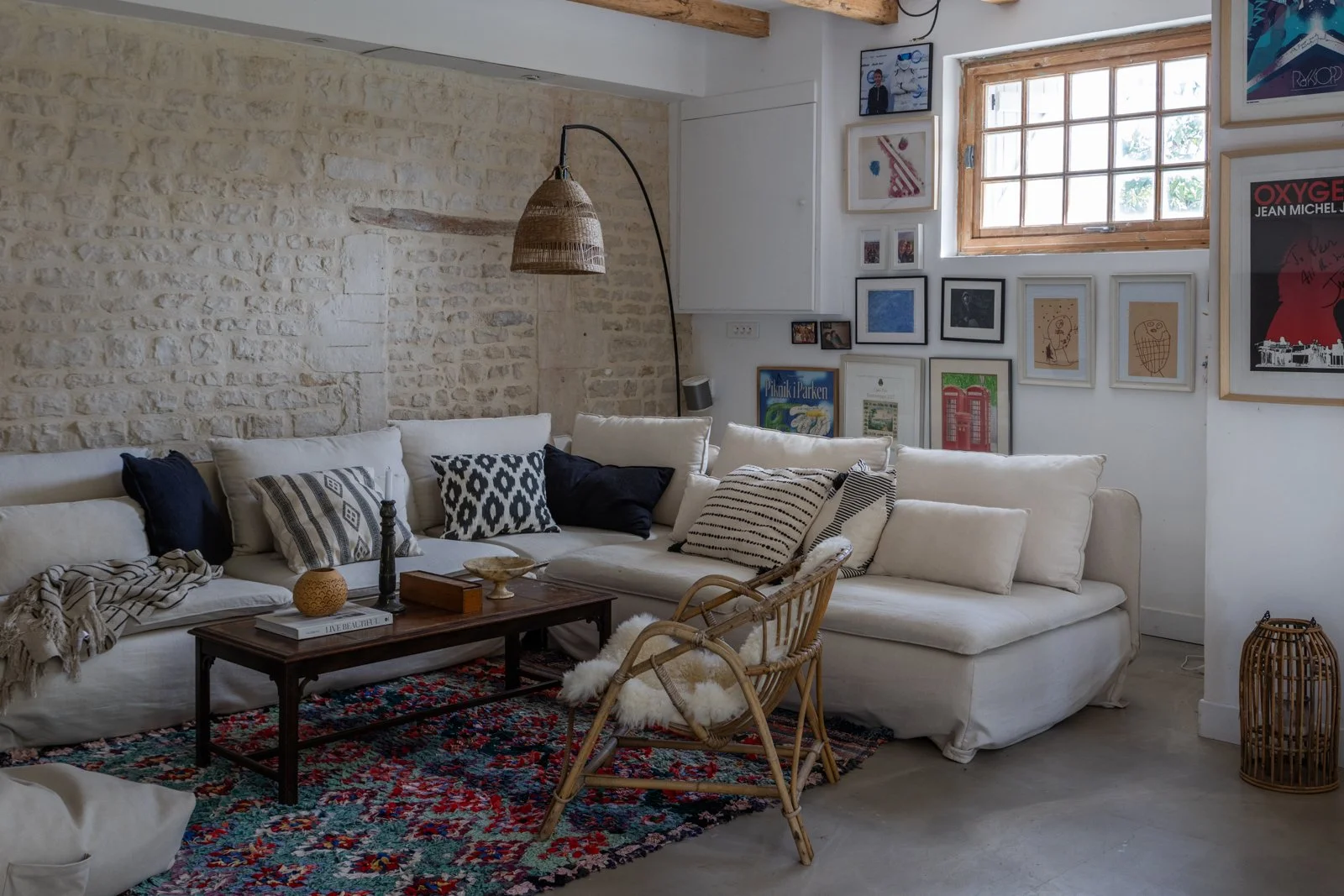 Cozy living room with white sectional sofa, assorted black and white patterned pillows, a colorful area rug, a wooden coffee table with decorative items, a rattan chair with a sheepskin, a wall with framed art and photos, a large window with wooden f