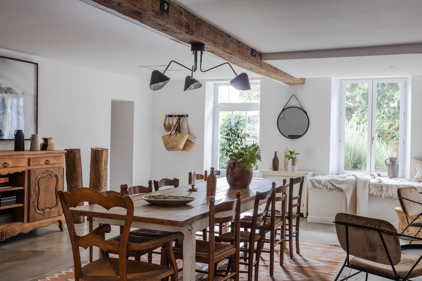 A rustic dining room with a wooden table, surrounded by assorted wooden chairs, situated in front of large windows letting in natural light. The room features a wooden beam across the ceiling, a modern black multi-light fixture, and decorative object