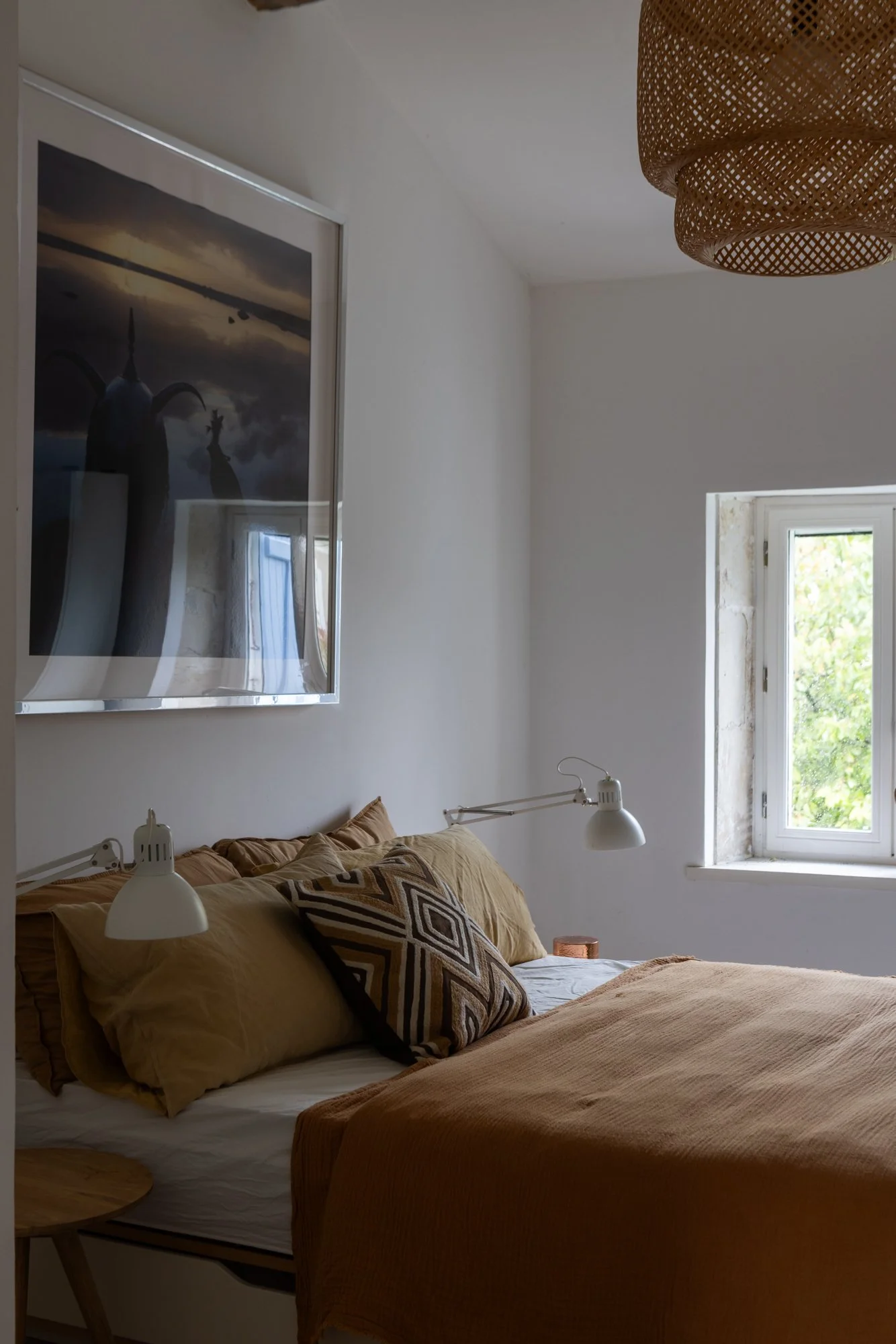 A cozy bedroom with a bed covered in beige and brown bedding, featuring patterned and solid-colored pillows. Two white wall-mounted lamps are on either side of the bed. A window on the right lets in natural light, and a large framed artwork hangs abo