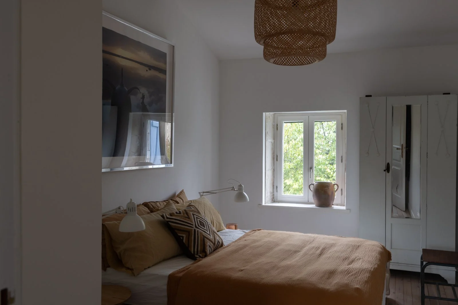 A bedroom with a bed dressed in tan bedding and decorative pillows, a window showing green trees outside, white walls, a wardrobe with a mirror, and a woven ceiling light fixture.